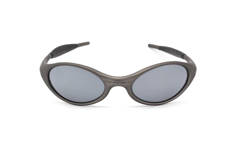 May include: Gray oval-shaped sunglasses with gray lenses. The sunglasses have a dark gray frame and temples. The brand name is subtly printed on the frame. A modern, sporty eyewear accessory.