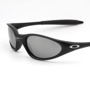 May include: Black sunglasses with gray lenses. The sunglasses have a curved frame and a small silver logo on the temple. The lenses are oval-shaped and the arms are thin and curved.