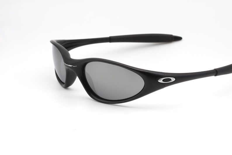 May include: Black sunglasses with gray lenses. The sunglasses have a curved frame and a small silver logo on the temple. The lenses are oval-shaped and the arms are thin and curved.