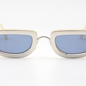 May include: A pair of silver-framed sunglasses with light blue lenses. The frames have a rounded, rectangular shape and are made of a smooth, shiny metal. The arms of the sunglasses are made of a cream-colored material.