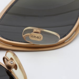 May include: Gold-toned sunglasses with a clear plastic temple tip that says "VERSACE" in gold lettering.