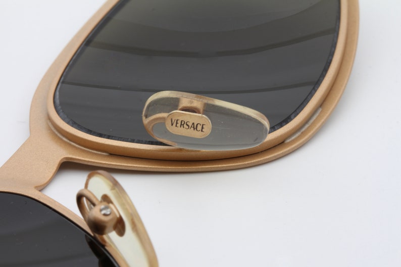 May include: Gold-toned sunglasses with a clear plastic temple tip that says "VERSACE" in gold lettering.