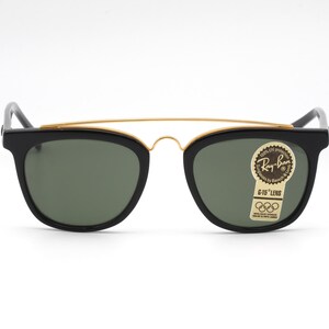 May include: Black Ray-Ban sunglasses with a gold-colored brow bar and green lenses. The sunglasses have a classic design with a rectangular frame and a sticker on the right lens. The sticker reads "Ray-Ban G-15 Lens".
