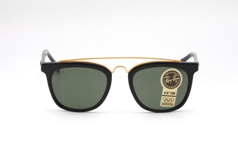 May include: Black Ray-Ban sunglasses with a gold-colored brow bar and green lenses. The sunglasses have a classic design with a rectangular frame and a sticker on the right lens. The sticker reads "Ray-Ban G-15 Lens".