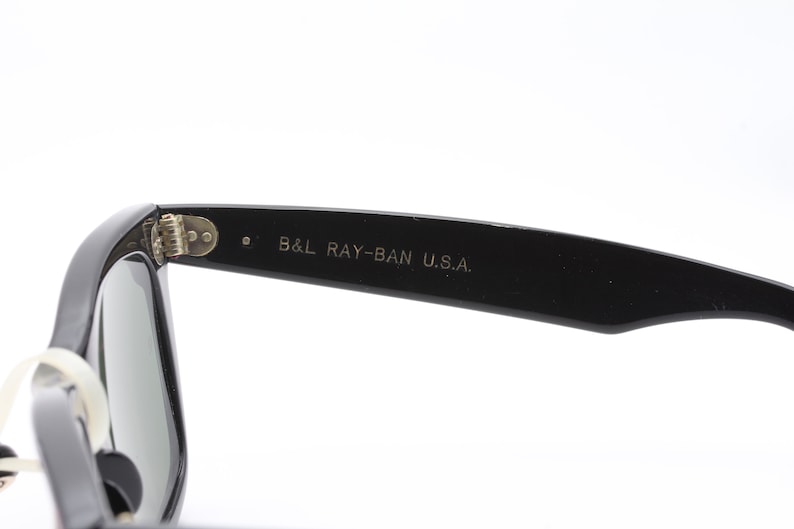 May include: Black Ray-Ban sunglasses with green lenses. The sunglasses have a metal hinge and the text "B&L RAY-BAN U.S.A." is printed on the arm.