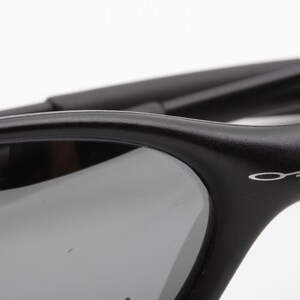May include: Close-up of black sunglasses with gray lenses. The frame is matte black with the brand name "Oakley" in white lettering on the temple. The lenses are a dark gray color, providing a contrast to the black frame.