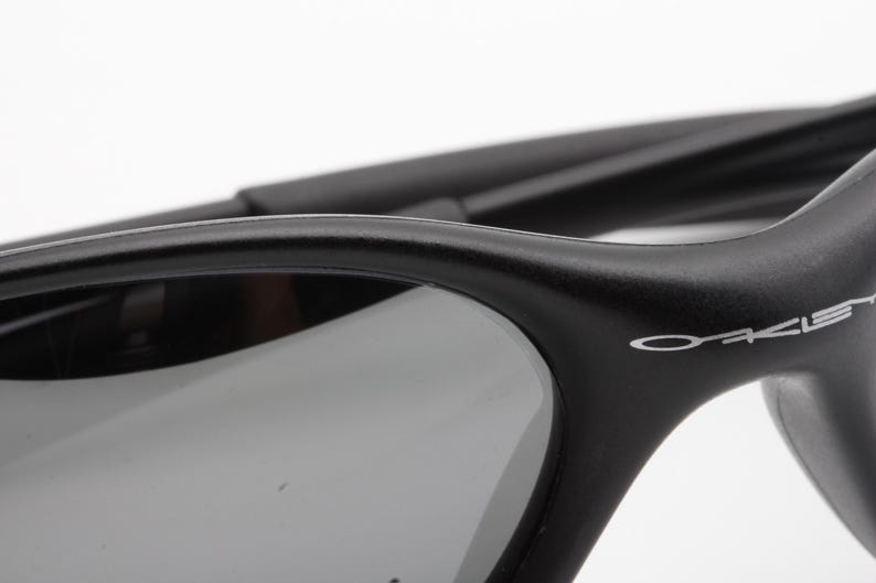 May include: Close-up of black sunglasses with gray lenses. The frame is matte black with the brand name "Oakley" in white lettering on the temple. The lenses are a dark gray color, providing a contrast to the black frame.
