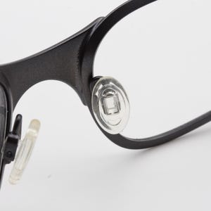 May include: Close-up of black eyeglasses with clear lenses. The glasses have a matte finish and feature clear nose pads for comfort. The frame is a simple, modern design.