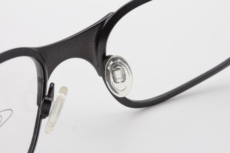 May include: Close-up of black eyeglasses with clear lenses. The glasses have a matte finish and feature clear nose pads for comfort. The frame is a simple, modern design.
