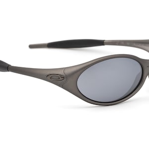 May include: Gray oval-shaped sunglasses with gray lenses. The sunglasses have a dark gray frame and arms, with a subtle logo on the side. The lenses are reflective, and the overall design is sleek and modern.