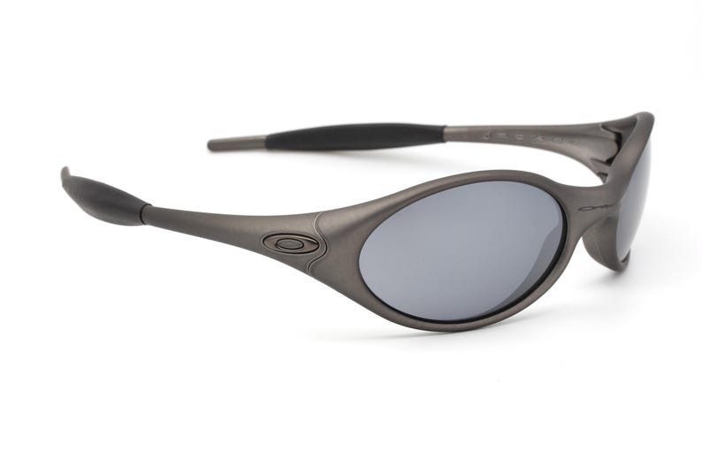 May include: Gray oval-shaped sunglasses with gray lenses. The sunglasses have a dark gray frame and arms, with a subtle logo on the side. The lenses are reflective, and the overall design is sleek and modern.