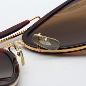 May include: Close-up of sunglasses with brown lenses and a dark brown frame. The frame has gold accents and a classic aviator style. The nose pads are clear and the temples are a darker brown.