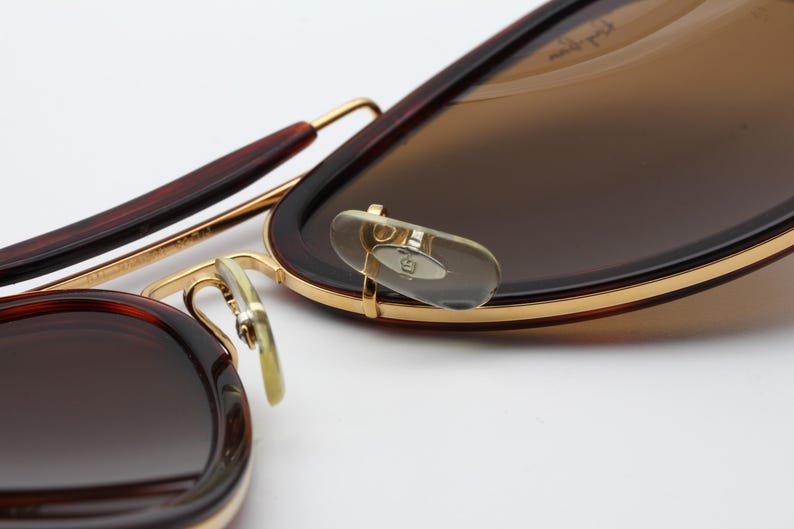 May include: Close-up of sunglasses with brown lenses and a dark brown frame. The frame has gold accents and a classic aviator style. The nose pads are clear and the temples are a darker brown.