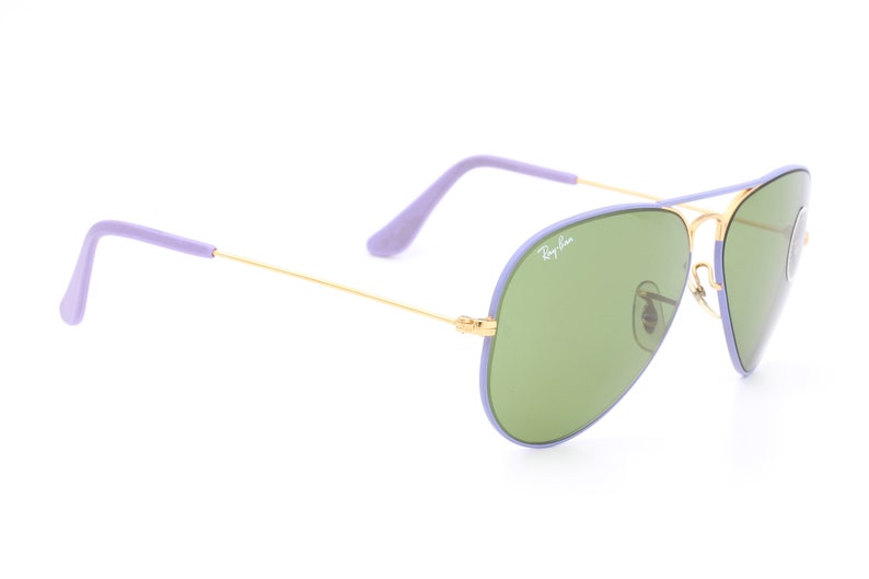 Op de afbeelding: Paarse en gouden aviator zonnebril met groene glazen. De zonnebril heeft een Ray-Ban logo op de rechter lens.