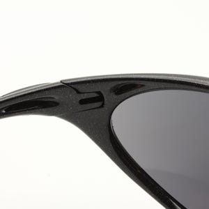 Puede incluir: Primer plano de gafas de sol negras con lentes grises oscuros. La montura tiene un aspecto texturizado y moteado. El dise&ntilde;o incluye una bisagra y una patilla curva con un recorte ovalado.