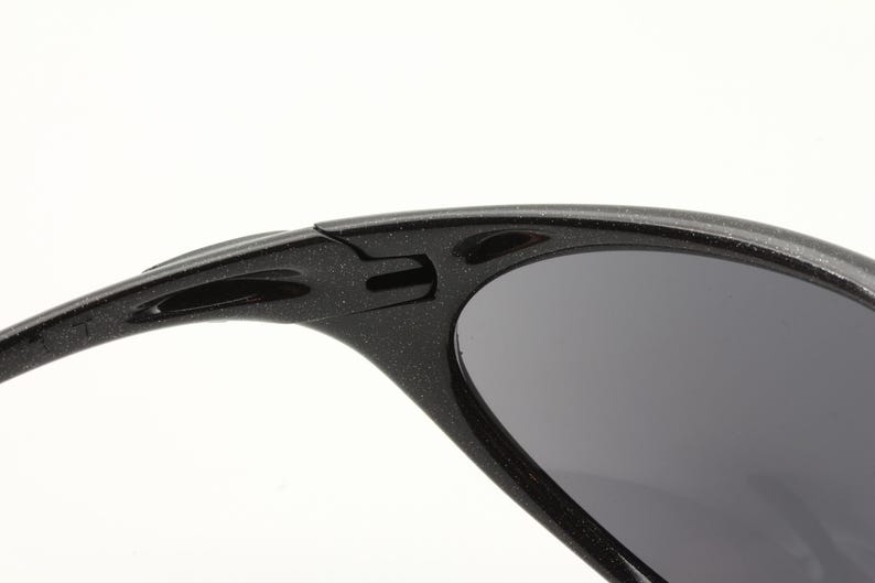 Puede incluir: Primer plano de gafas de sol negras con lentes grises oscuros. La montura tiene un aspecto texturizado y moteado. El dise&ntilde;o incluye una bisagra y una patilla curva con un recorte ovalado.