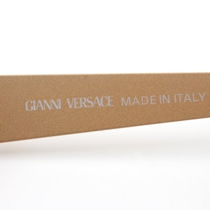 May include: Close-up of the gold-toned arm of a pair of sunglasses with the text "GIANNI VERSACE MADE IN ITALY" printed on the arm.
