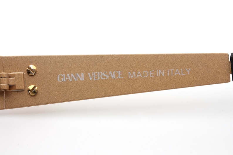 May include: Close-up of the gold-toned arm of a pair of sunglasses with the text "GIANNI VERSACE MADE IN ITALY" printed on the arm.