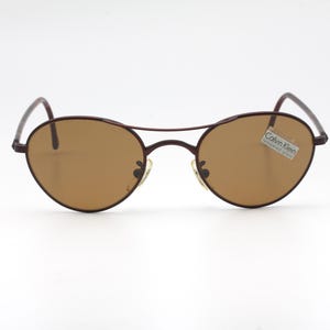May include: Brown metal frame sunglasses with brown lenses. The sunglasses have a Calvin Klein label on the right lens.