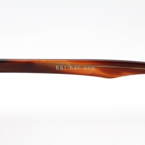 May include: Close-up of the brown tortoise shell frame of a pair of Ray-Ban sunglasses. The frame is engraved with the text "B&L RAY-BAN".