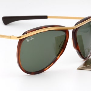 Pu&ograve; includere: Un paio di occhiali da sole aviatore Ray-Ban con montatura tartaruga e dettagli dorati. Le lenti sono verdi e gli occhiali sono nuovi con etichette.
