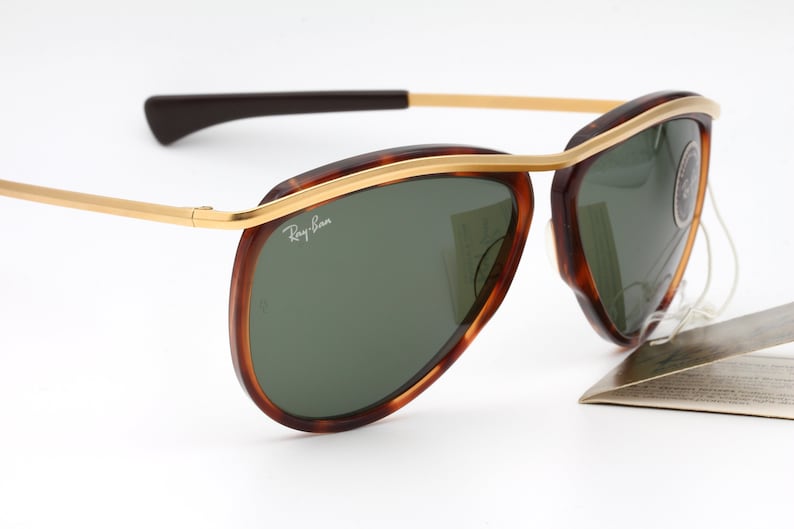Pu&ograve; includere: Un paio di occhiali da sole aviatore Ray-Ban con montatura tartaruga e dettagli dorati. Le lenti sono verdi e gli occhiali sono nuovi con etichette.