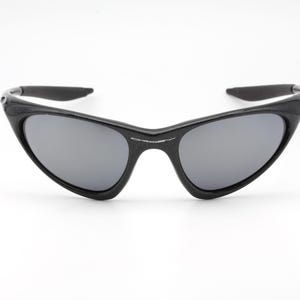 Puede incluir: Gafas de sol negras con lentes grises. Las gafas tienen un dise&ntilde;o elegante y angular con un acabado mate. Las monturas son gruesas y envuelven las lentes, proporcionando un aspecto moderno y deportivo. Las patillas est&aacute;n ligeramente curvadas.