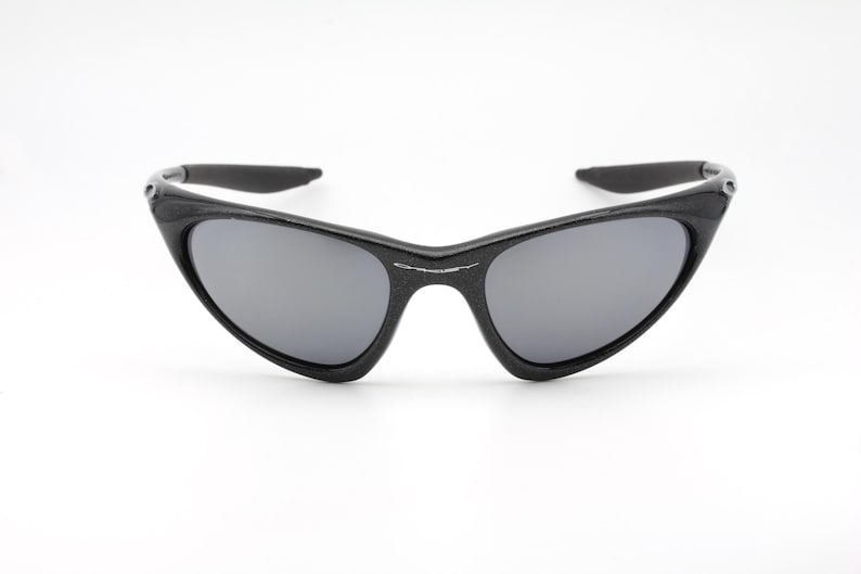 Puede incluir: Gafas de sol negras con lentes grises. Las gafas tienen un dise&ntilde;o elegante y angular con un acabado mate. Las monturas son gruesas y envuelven las lentes, proporcionando un aspecto moderno y deportivo. Las patillas est&aacute;n ligeramente curvadas.