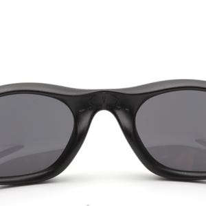 May include: Close-up of black sunglasses with dark gray lenses. The sunglasses have a curved, streamlined design and a matte finish. The lenses are oval-shaped and provide a contrast to the black frame.