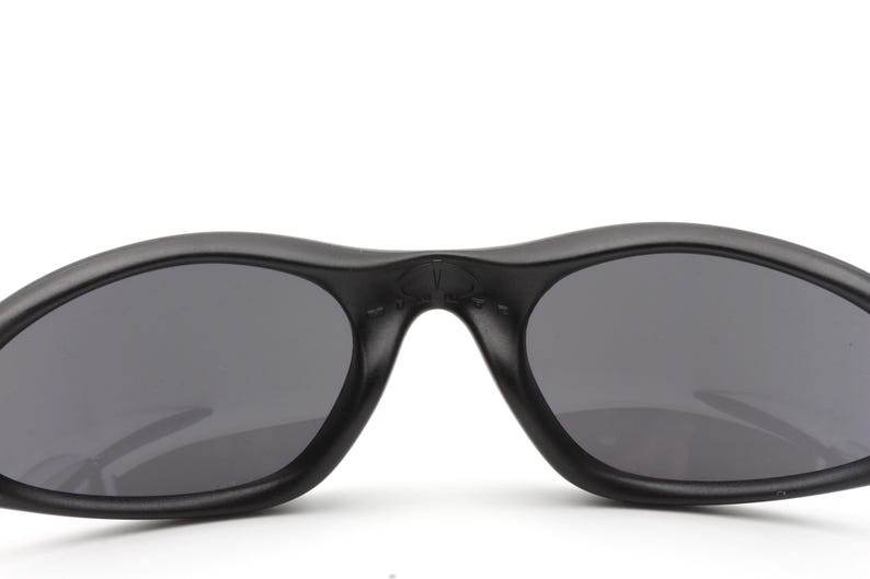 May include: Close-up of black sunglasses with dark gray lenses. The sunglasses have a curved, streamlined design and a matte finish. The lenses are oval-shaped and provide a contrast to the black frame.