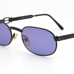 Può includere: Occhiali da sole ovali neri con cinturino in pelle e fibbia in metallo. Le lenti sono di colore viola scuro.