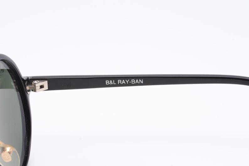 May include: Black Ray-Ban aviator sunglasses with green lenses. The sunglasses have a metal frame and the text "B&L RAY-BAN" is printed on the frame.