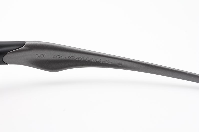 Może przedstawiać: Zbliżenie ciemnoszarego zausznika okular&oacute;w. Na zauszniku wydrukowano napis "CE MADE IN U.S.A.". Zausznik ma elegancki, zakrzywiony kształt.