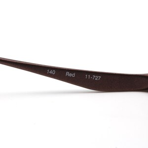 May include: Close-up of the brown metal arm of a pair of glasses. The arm is engraved with the text "140 Red 11-727".