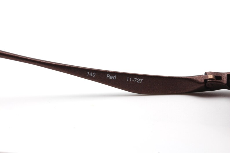 May include: Close-up of the brown metal arm of a pair of glasses. The arm is engraved with the text "140 Red 11-727".