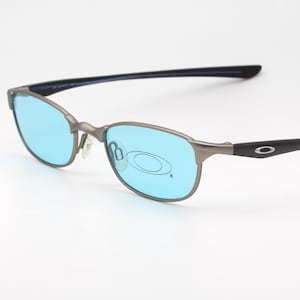 Puede incluir: Gafas de sol con montura metálica gris y lentes azul claro. Las patillas son negras con un logotipo plateado. Las gafas tienen un diseño elegante y moderno, adecuado para actividades al aire libre o uso diario.