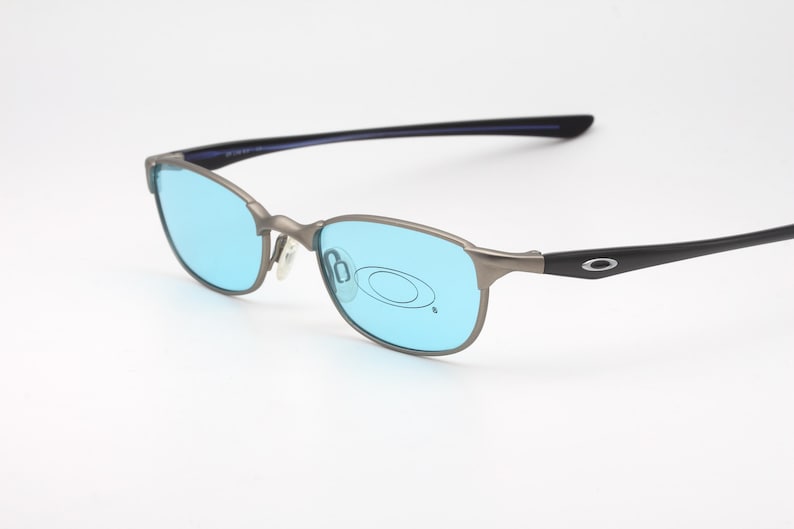 May include: Gray metal-framed sunglasses with light blue lenses. The temples are black with a silver logo. The glasses have a sleek, modern design, suitable for outdoor activities or everyday wear.