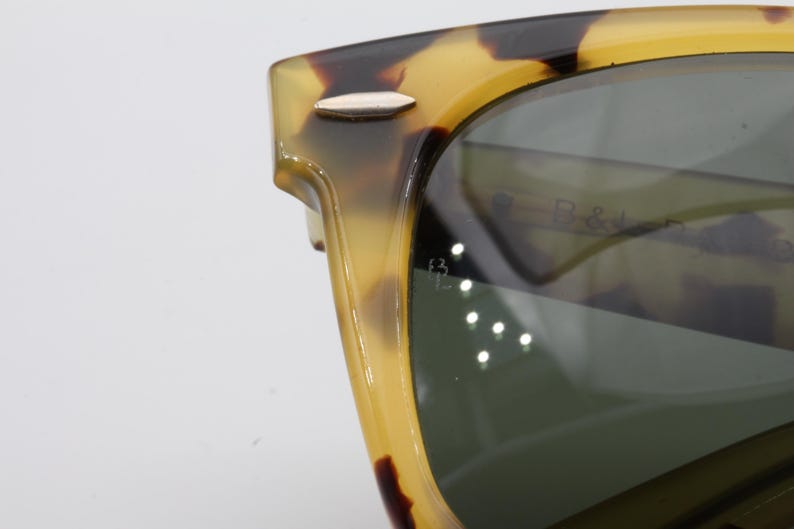 Puede incluir: Primer plano de gafas de sol con montura con estampado de carey. Las lentes son de color verde oscuro y la montura tiene un detalle plateado en la patilla. La marca "B & J" es visible en la lente.