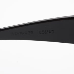 May include: Close-up of a black Wayfarer Nomad sunglasses arm. The text "WAYFARER NOMAD" is embossed on the arm. The sunglasses are set against a white background.