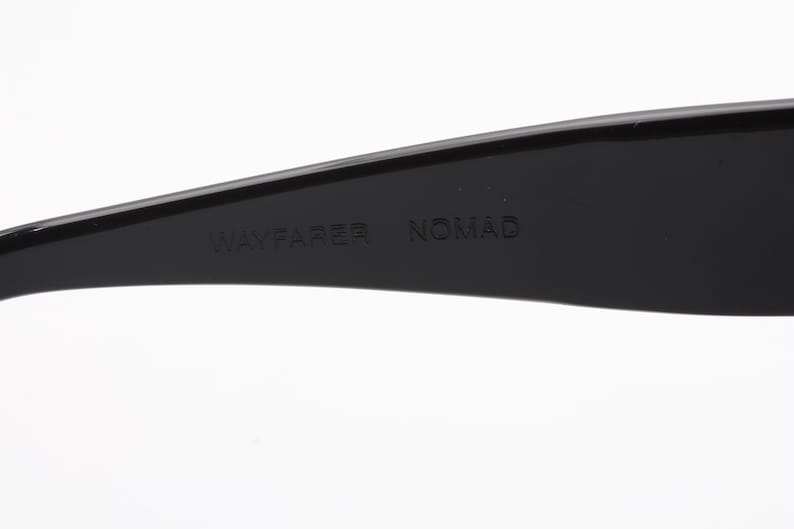 May include: Close-up of a black Wayfarer Nomad sunglasses arm. The text "WAYFARER NOMAD" is embossed on the arm. The sunglasses are set against a white background.