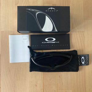 May include: Black Oakley sunglasses with a black case and box. The box has the word "MINUTE" printed on it. The sunglasses are in a black soft case. A white warranty card and a "STAMP" card are also present.