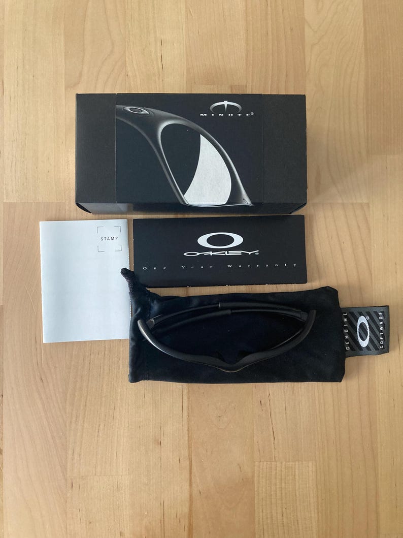 May include: Black Oakley sunglasses with a black case and box. The box has the word "MINUTE" printed on it. The sunglasses are in a black soft case. A white warranty card and a "STAMP" card are also present.
