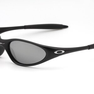 May include: Black sunglasses with gray lenses. The sunglasses have a curved, wraparound design with a matte finish. The Oakley logo is visible on the temples. The sunglasses are set against a white background.