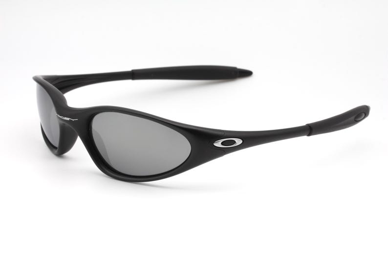 May include: Black sunglasses with gray lenses. The sunglasses have a curved, wraparound design with a matte finish. The Oakley logo is visible on the temples. The sunglasses are set against a white background.