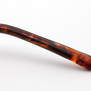 May include: Close-up of a portion of eyeglasses. The arm of the glasses is a mottled brown and amber color, with a curved design. The end of the arm is rounded. The background is white.