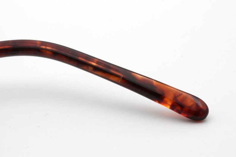 May include: Close-up of a portion of eyeglasses. The arm of the glasses is a mottled brown and amber color, with a curved design. The end of the arm is rounded. The background is white.
