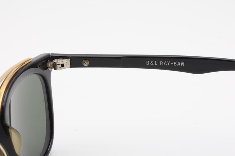 May include: Black Ray-Ban sunglasses with gold accents. The sunglasses have a classic design with a rectangular frame and green lenses. The text "B&L RAY-BAN" is printed on the temple.