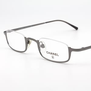 Puede incluir: Gafas con montura metálica gris con forma de lente rectangular. El logotipo de Chanel es visible en la lente derecha. Las gafas tienen un diseño minimalista con montura y varillas finas.