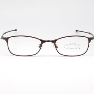 Puede incluir: Un par de gafas de metal marrón con lentes transparentes. Las lentes tienen una pequeña forma ovalada en el centro.