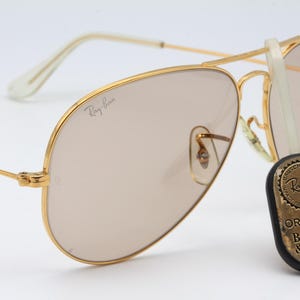 May include: Gold-tone aviator sunglasses with brown lenses and a Ray-Ban logo on the frame. The sunglasses are accompanied by a round, gold-tone metal tag with the text "Ray-Ban Original Bausch & Lomb".